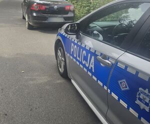 Na zdjęciu widać bok radiowozu policyjnego i innego samochodu osobowego. Oba pojazdy są zaparkowane przy drodze.