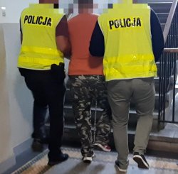 Na zdjęciu nieumundurowani policjanci prowadzą skutego w kajdanki zatrzymanego mężczyznę po schodach w budynku komendy.