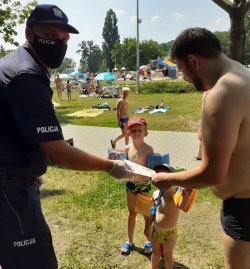Policjant w maseczce przekazuje plażowiczowi ulotki na temat bezpieczeństwa, w tle biegające dzieci
