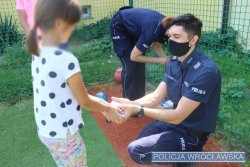 Policjant wręcza dziewczynce upominek, w tle policjantka wyciąga kolejne upominki dla pozostałych dzieci