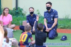Policjant i policjantka siedzą wspólnie z dziećmi na boisku, jedno z dzieci trzyma podniesioną do góry rękę