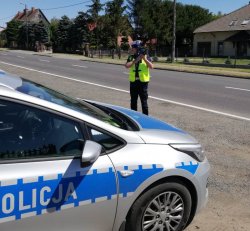 Policjant stojący przed radiowozem na drodze mierzy prędkość nadjeżdżającym pojazdom