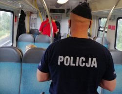 Umundurowany policjant stojący tyłem, w pociągu Kolei Dolnośląskich kontroluje pod kątem przestrzegania nowych obostrzeń, czy pasażerowie mają założone maseczki i zachowują odstęp. W tle policjant rozmawiający z kierownikiem pociągu.