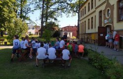 Na zdjęciu dzieci siedzące na ławkach i ławeczkach przed budynkiem, odwrócone tyłem do obiektywu, skierowane wzrokiem w kierunku policjantki która mówi o bezpieczeństwie podczas wakacji w Krośnicach 21.08.2020 r.