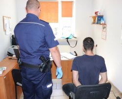 Na zdjęciu policjant z zatrzymanym w trakcie przedstawiania mu zarzutów. Policjant stoi obok siedzącego na krześle zatrzymanego mężczyzny.