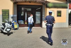 Na zdjęciu policjant zmierzający do drzwi placówki handlowej, przed nim znajduje się mężczyzna w maseczce ochronnej.