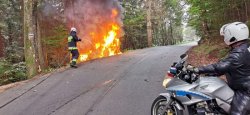 Na zdjęciu na pierwszym planie policjant na motorze w tle widoczna droga i strażak gaszący w rowie palący się pojazd marki Audi