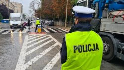 Policjanci pilnują bezpieczeństwa na  przejściu dla pieszych, zatrzymując samochody i przepuszczając pieszych.