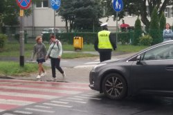 Na zdjęciu policjant stojący przy pasach dla pieszych, dwie kobiety przechodzące przez pasy i stojący przed pasami samochód