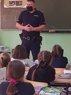 Na zdjęciu widać na środku stojącego policjanta w mundurze, który przemawia do dzieci siedzących w ławce. Zdjęcie przedstawia klasę szkolną.