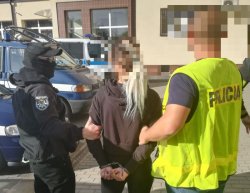 Policjant i policjantka prowadzą zatrzymaną kobietę. przez parking policyjny. Kobieta ma na rękach kajdanki.