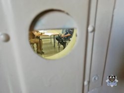 Zdjęcie przez wizjer w drzwiach przez który widać policjanta i zatrzymanego siedzących przy stole podczas przesłuchania.