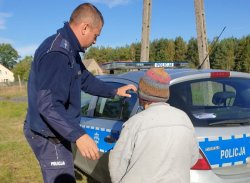 Na zdjęciu policjant pomagającej odnalezionej starszej kobiecie wsiąść do radiowozu, aby mógł odwieźć ją do domu.
