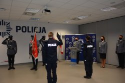 Na zdjęciu widać tyłem stojących dwóch policjantów mających podniesioną prawą rękę do góry, za nimi stoi trzech policjantów trzymających sztandar jednostki. W tle widać lektora oraz pozostałych policjantów. Na ścianie widnieje napis policja.