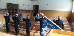 Na zdjęciu widać salę z dużą ilością krzeseł a w niej, w pierwszym rzędzie, policjantów umundurowanych z podniesioną prawą ręką do góry i wysuniętymi dwoma palcami. Recytują słowa Roty. Widać również sztandar jednostki oraz dwóch policjantów stojących w drugim rzędzie.