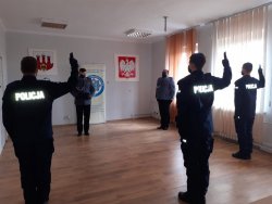 Na zdjęciu widać policjantów stojących tyłem z podniesioną prawą dłonią do góry, z wysuniętymi dwoma palcami. Powtarzają słowa roty ślubowania wypowiadane przez lektora widocznego w tle zdjęcia. Po prawej stronie widać również stojącego na baczność kierownika jednostki. Na ścianie wisi godło oraz herb powiatu.