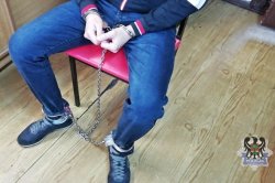 Na zdjęciu zatrzymany mężczyzna siedzi na krześle z kajdankami zespolonymi na rękach i nogach.