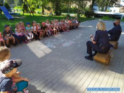 Na zdjęciu na dworze siedzi dwoje policjantów, a przed nimi dzieci. Słuchają co opowiadają policjanci. W tle widać plac zabaw.