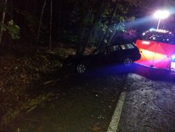 Zdjęcie zrobione w porze nocnej, pojazdu, który uderzył w drzewo. W tle widoczne światła błyskowe Straży Pożarnej.