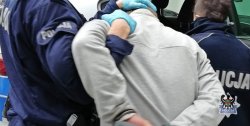 Na zdjęciu dwóch umundurowanych policjantów prowadzi zatrzymanego mężczyznę, który na rękach ma kajdanki.