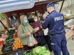 Na zdjęciu kiosk z warzywami stojący przy drodze  i kobiety robiące w nim zakupy, na pierwszym planie policjant przekazujący jednej z kupujących ulotkę informacyjną. Wszyscy mają maseczki ochronne na twarzy.