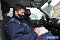 Na zdjęciu policjant oraz policjantka siedzą w radiowozie. Na twarzach mają maseczki ochronne. Policjantka przegląda dokumenty.