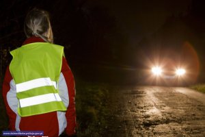 Na zdjęciu nocną porą kobieta w kamizelce odblaskowej idąca lewą stroną pobocza, a naprzeciwko nadjeżdża samochód.
