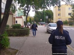 Na zdjęciu widać umundurowaną policjantkę, która idzie po chodniku, a w tle widać mężczyznę oraz jeden z bloków mieszkalnych i zaparkowany pojazd.