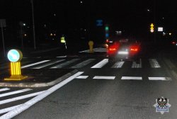 Na zdjęciu nocna porą, przedstawione jest miejsce wypadku drogowego, w tym samochód, który potrącił pieszego oraz policjanta na miejscu zdarzenia nieopodal przejścia dla pieszych.