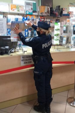 Na zdjęciu policjantka przykleja na tablicę ogłoszeń w aptece materiały prewencyjne dotyczące bezpieczeństwa seniorów.