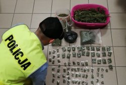 Na zdjęciu policjant pochylający się nad zabezpieczonymi narkotykami: marihuaną i amfetaminą w woreczkach foliowych i pojemnikach plastikowych.