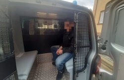 Na zdjęciu zatrzymany mężczyzna siedzący w kajdankach, w radiowozie policyjnym.