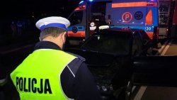 Na zdjęciu na pierwszym planie policjant stojący nad zniszczonym samochodem, który brał udział w kolizji oraz w dali karetka pogotowia.