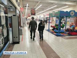 Na zdjęciu policjantka i żołnierz idą korytarzem galerii handlowej po lewej stronie ciąg sklepów a po prawej karuzela dla dzieci.