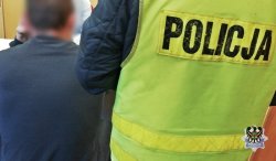 Zdjęcie przedstawia zatrzymana osobę i stojącego obok tyłem policjanta w odblaskowej kamizelce z napisem policja