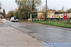 Na zdjęciu widać przejazd kolejowy i drogę asfaltową oraz znak STOP. W oddali znajdują się budynki i przejeżdżający samochód.