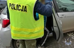 Na zdjęciu widać funkcjonariusza w kamizelce odblaskowej z napisem POLICJA, który pomaga wsiąść zatrzymanemu mężczyźnie do radiowozu policyjnego.