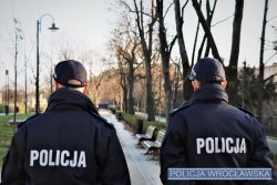 Na zdjęciu widać dwóch umundurowanych policjantów od tyłu, a w tle drzewa i droga dla rowerów oraz fosa miejska.