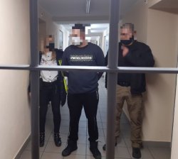 Na zdjęciu widok przez kraty zatrzymanego mężczyzny w kajdankach oraz prowadzącego go do celi policjantki i policjanta w ubraniu cywilnym.