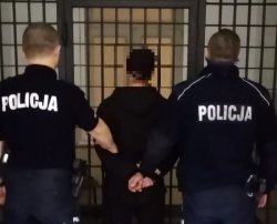 Na zdjęciu dwóch policjantów prowadzi zatrzymanego mężczyznę do celi. W tle widać kraty.
