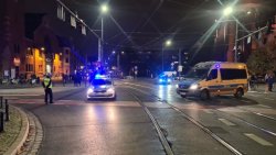 Skrzyżowanie na którym stoi umundurowany policjant w kamizelce odblaskowej koloru żółtego z czarnym napisem POLICJA. Na skrzyżowaniu są też dwa radiowozy policyjne oznakowane oraz jeden nieoznakowany. Wszystkie pojazdy mają włączone sygnały błyskowe barwy niebieskiej.