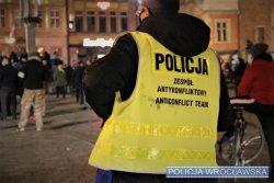 Funkcjonariusz policji w żółtej kamizelce odblaskowej z czarnym napisem POLICJA zespół antykonfliktowy anticonflict team. W tle osoby postronne i pręgierz rynku wrocławskiego.