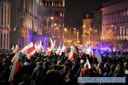 Marsz 11 listopada ulicami Wrocławia. Uczestnicy mają flagi Polski i symbole narodowe. W tle kamienice i miejskie zabudowania oraz radiowozy policyjne z włączonymi sygnałami świetlnymi barwy niebieskiej.