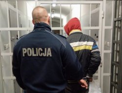 Na zdjęciu policjant prowadzony zatrzymanego mężczyznę w kapturze z kajdankami na rękach do aresztu. W tle widać kraty.
