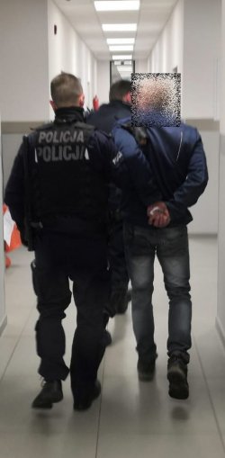 Zdjęcie przedstawia dwóch policjantów, jeden z nich prowadzi zatrzymanego mężczyznę, który ma założone kajdanki na ręce trzymane z tyłu. Idą korytarzem pomiędzy pomieszczeniami dla osób zatrzymanych