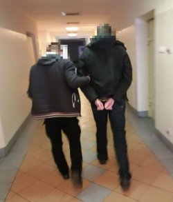 Na zdjęciu policjant w cywilnym ubraniu prowadzi skutego w kajdanki zatrzymanego korytarzami komendy policji.