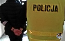 Na zdjęciu policjant prowadzi skutego w kajdanki zatrzymanego mężczyznę.