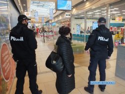 Zdjęcie przedstawia patrol umundurowanych policjantów znajdujących się w holu galerii handlowej. Pomiędzy policjantami stoi kobieta. W tle widać witryny sklepowe.