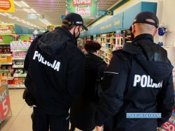Zdjęcie przedstawia dwóch policjantów zaglądających do sklepu. Stoją w holu galerii handlowej, wraz z nimi znajduje się pracownik sanepidu. W tle widać witryny sklepowe.