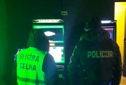 Na zdjęciu nieumundurowany funkcjonariusz Kontroli Celno-Skarbowej z ubrany w kamizelkę odblaskową z napisem służba celna i nieumundurowany policjant w kamizelce z napisem policja stoją przed dwoma automatami do gier.